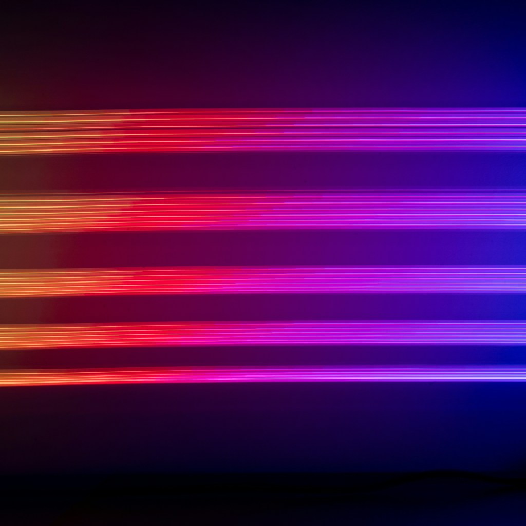 Neon lights, five in total appear to be on a black ceiling. They are in hues of (from left to right). yellow, pink, red, purple, blue. Photo by Adrien via unsplash.com